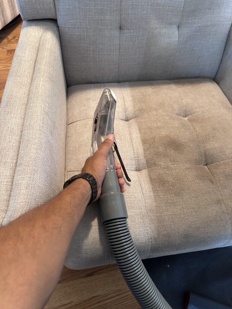 Upholstery cleaning tool on a gray tufted armchair, with the cleaned section of the seat cushion visibly lighter than the soiled fabric beside it